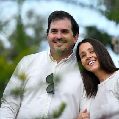 A smiling man and woman stand close together in a natural outdoor setting.