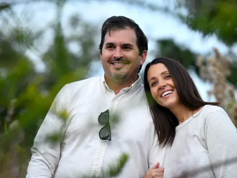 A smiling man and woman stand close together in a natural outdoor setting.