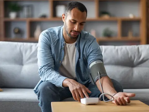Young Man Sitting On Couch And Checking His Blood Pressure With Upper Arm Monitor.