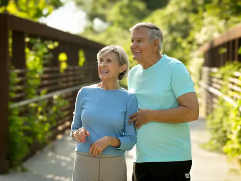 Smiling older couple in casual athletic wear walking arm-in-arm on a wooden bridge in a sunny outdoor park setting