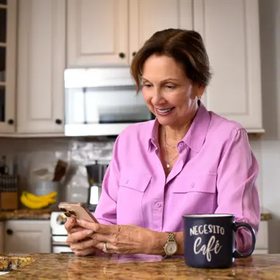 A smiling woman in a pink shirt uses her smartphone in a modern kitchen with a coffee mug nearby.