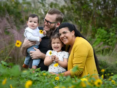 A family of four, two adults and two children, smiling and holding flowers in a field.