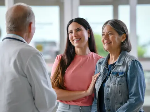 A smiling doctor talks to two smiling women in a hospital setting.