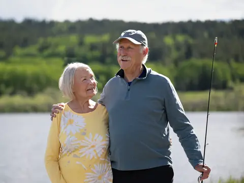 Elderly couple standing by a lake, smiling and holding fishing rods.