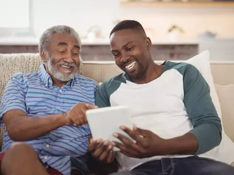 Two men sitting on a couch, smiling and looking at a tablet.