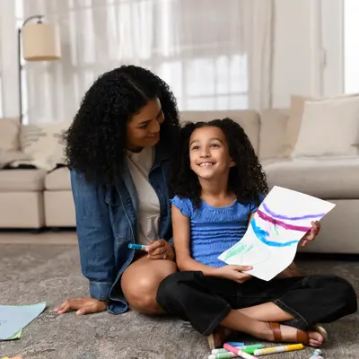 Mother and daughter sitting on living room floor, child holding up colorful drawing while mother smiles at her, with art supplies nearby