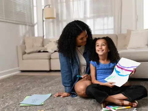 Mother and daughter sitting on living room floor, child holding up colorful drawing while mother smiles at her, with art supplies nearby