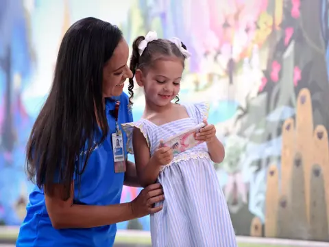 A smiling woman in blue scrubs helps a young girl in a striped dress, both looking at a book.