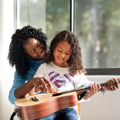 Mother and daughter smiling while playing a guitar together at home.