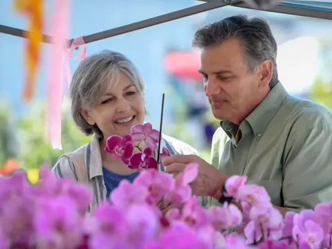 A man and woman are smiling and looking at purple orchids at a flower stand.