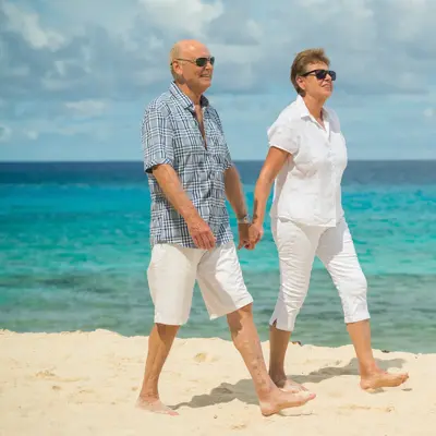 Older couple in casual white clothing walking hand-in-hand on a sandy beach with turquoise ocean water in the background