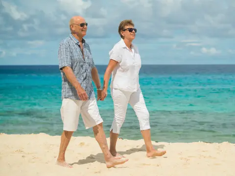 Older couple in casual white clothing walking hand-in-hand on a sandy beach with turquoise ocean water in the background