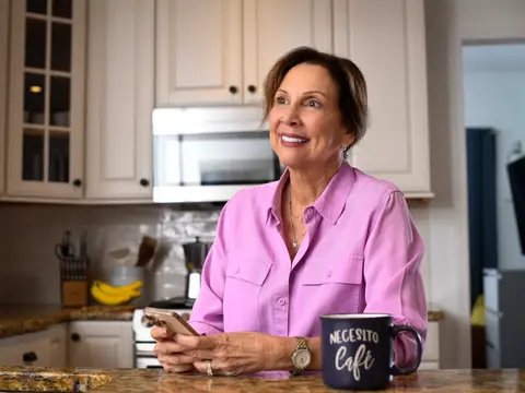 A smiling woman in a pink shirt sits at a kitchen counter with a smartphone and coffee mug.