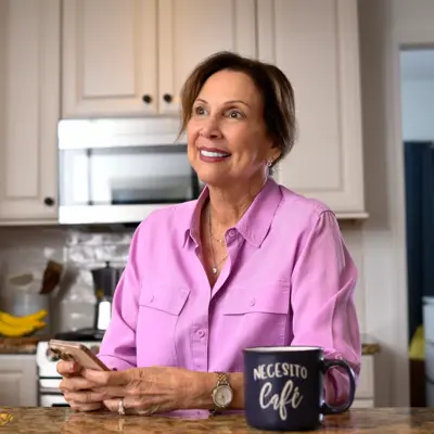 A smiling woman in a pink shirt sits at a kitchen counter with a smartphone and coffee mug.