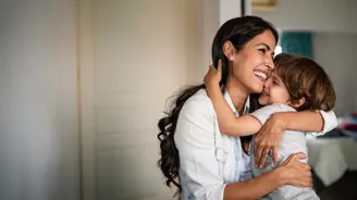 Smiling adult and young child share a warm hug indoors, faces pressed together in an affectionate embrace.