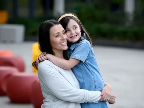 A smiling woman hugs a young girl in a park, both wearing casual clothing.