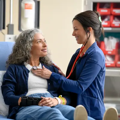Healthcare professional in blue scrubs using a stethoscope to examine a smiling patient seated in a blue chair in a modern medical facility