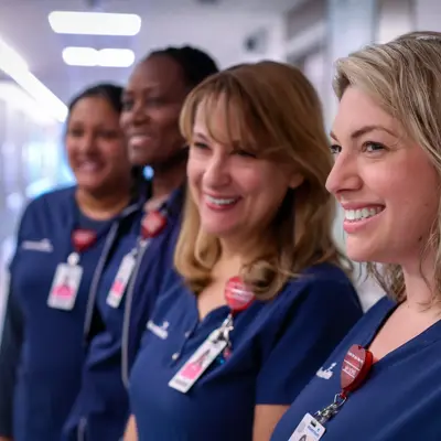 Four smiling nurses in blue uniforms stand together in a hospital corridor.