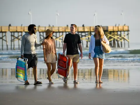 Group of four friends walking on the beach with beach chairs and a bag, with a pier in the background.