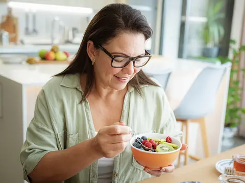 Woman wearing glasses and a green shirt smiling while holding an orange bowl filled with fresh fruit including strawberries, blueberries, and kiwi in a bright modern kitchen