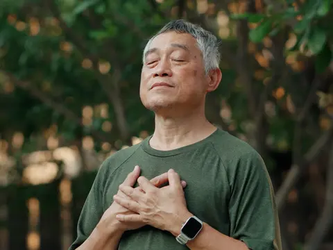 An older man with eyes closed and hands placed over his heart, practicing deep breathing exercises outdoors in a natural setting with trees in the background