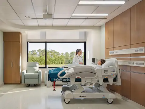A modern medical-surgical patient room with a patient in a hospital bed attended by a healthcare provider in a white coat, featuring a large window with a natural view, light blue accent wall, wood cabinetry, and a comfortable guest chair