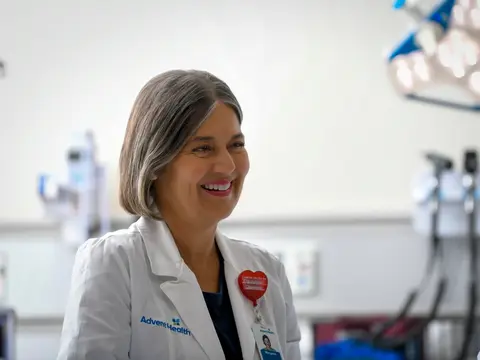 A smiling female doctor in a white lab coat with AdventHealth logo stands in a hospital room.