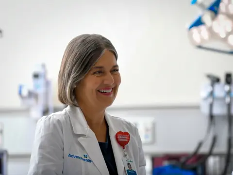A smiling female doctor in a white lab coat with AdventHealth logo stands in a hospital room.