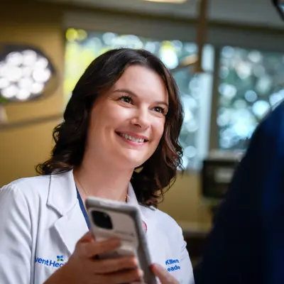A smiling female doctor in a white coat holds a smartphone, engaging with a patient.