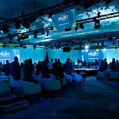 A large conference room with many attendees, blue lighting, and screens displaying 'At the Heart'.