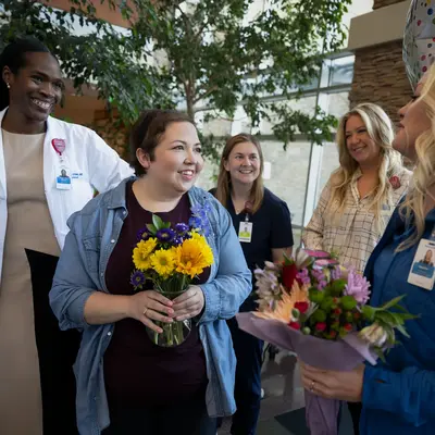 Group of smiling women in a hospital setting, one holding flowers, celebrating together.