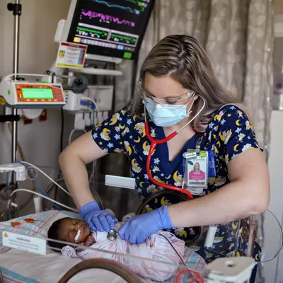Nurse in blue scrubs and face mask using a stethoscope on a newborn in an incubator.