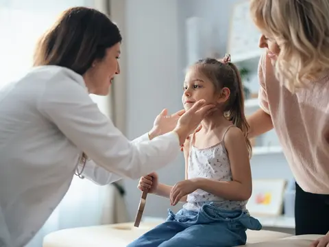 Pediatrician in white coat examining a young girl's face while her mother stands beside them in a bright clinic room