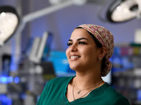 Smiling healthcare professional in green scrubs and floral surgical cap standing in a surgical suite with medical equipment visible in the background