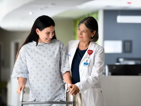 A smiling patient with a walker is supported by a smiling doctor in a hospital corridor.