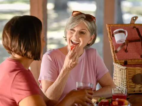 Two women enjoying a picnic, one eating a strawberry and smiling.