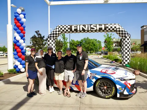 Six team members in matching black polo shirts pose beside a branded NASCAR race car beneath a checkered finish arch decorated with red, white, and blue balloon columns at an outdoor event.