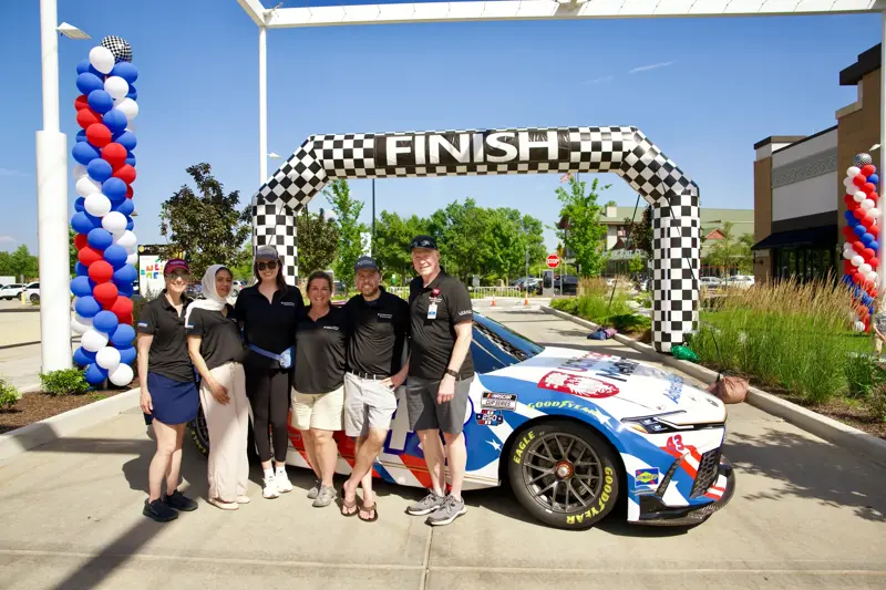 Six team members in matching black polo shirts pose beside a branded NASCAR race car beneath a checkered finish arch decorated with red, white, and blue balloon columns at an outdoor event.