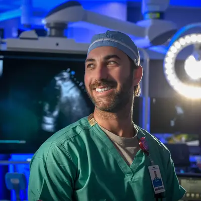 Smiling male surgeon in green scrubs stands in a modern operating room with medical equipment.