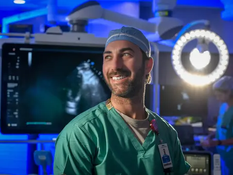 Smiling male surgeon in green scrubs stands in a modern operating room with medical equipment.