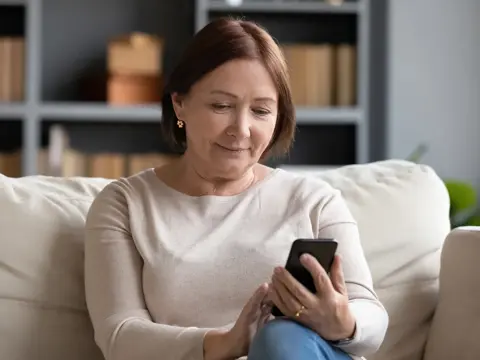 Middle-aged woman in a beige sweater sitting on a couch looking down at her smartphone