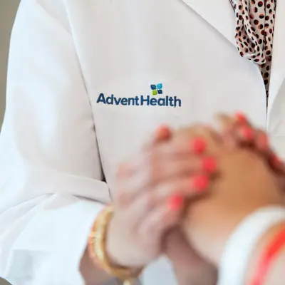 A close-up of a doctor and patient holding hands, with the AdventHealth logo visible on the doctor's coat.
