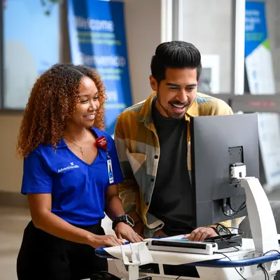 A smiling AdventHealth employee helps a patient at a computer kiosk in a hospital.