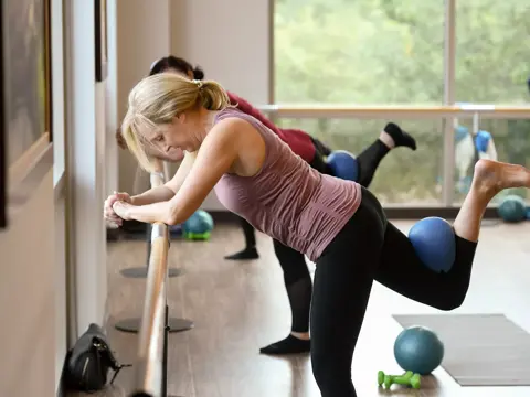 A woman in a pink tank top and black leggings performs a leg exercise using a blue exercise ball in a gym.