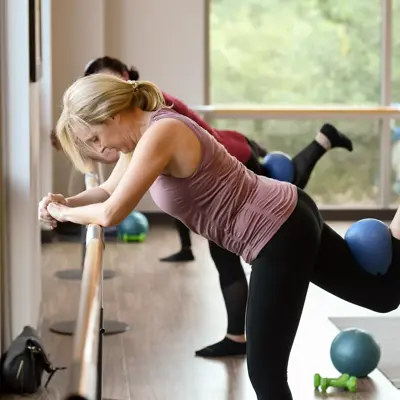 A woman in a pink tank top and black leggings performs a leg exercise using a blue exercise ball in a gym.