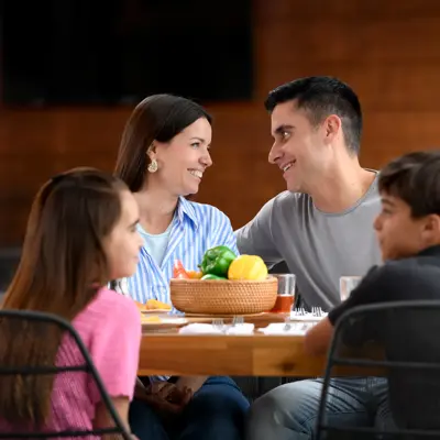 Hispanic family of four sitting together at a dining table with a bowl of fresh vegetables, smiling and enjoying a meal in a warm home setting.
