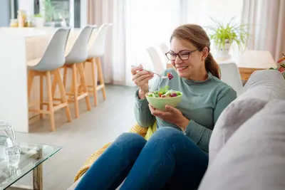 A woman sitting on a couch eats from a bowl of salad.