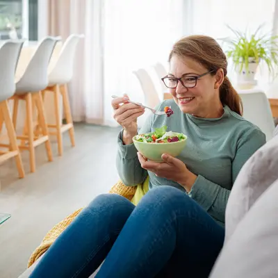 A woman sitting on a couch eats from a bowl of salad.