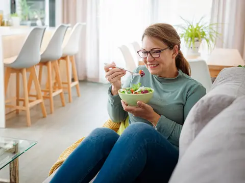 A woman sitting on a couch eats from a bowl of salad.