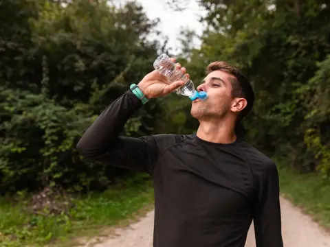 Man in black long-sleeve shirt drinks water from a plastic bottle while standing on a forest trail.